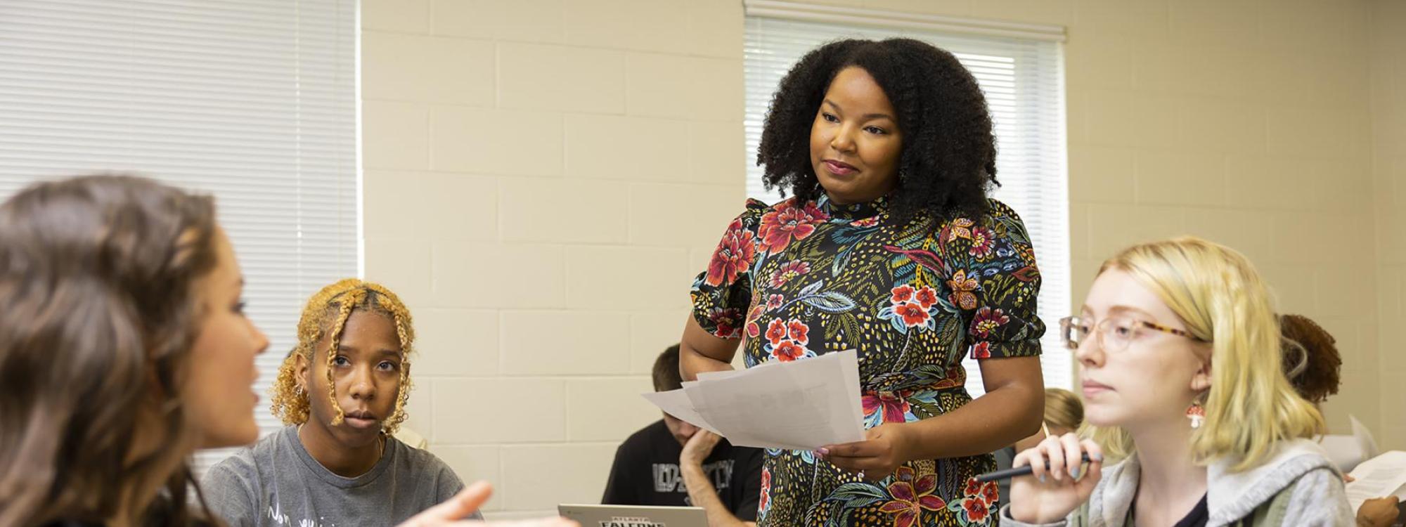 Dr. Johnson standing in her class amongst her students who are all sitting at their desks in a circle while one student has an active discussion with her. 